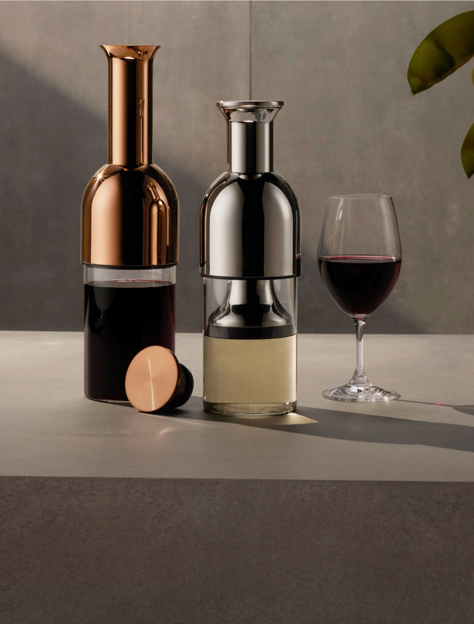 A copper mirror eto wine decanter with red wine in it beside a stainess mirror wine decanter with white wine in it, beside a glass of red wine