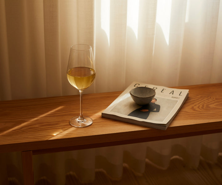A glass of white wine on wooden bench beside a magazine with a small bowl on it